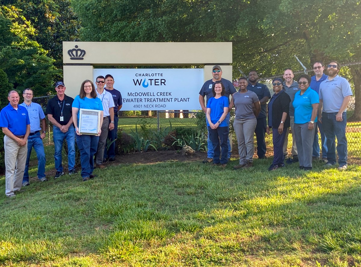 15 utility employees and supervisors line up on grass to pose for group photo in front of a large sign Charlotte Water McDowell Creek Wastewater Treatment Plant 4901 Neck Road