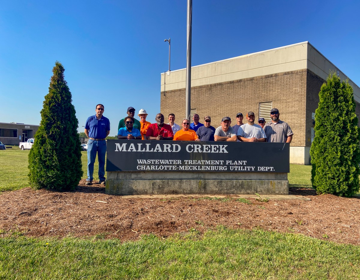 14 utility employees and supervisors pose for a group photo standing behind a charcoal grey stone sign Mallard Creek Wastewater Treatment Plant Charlotte Mechlenburgn Utility Dept, and a one-story brick building appears behind them