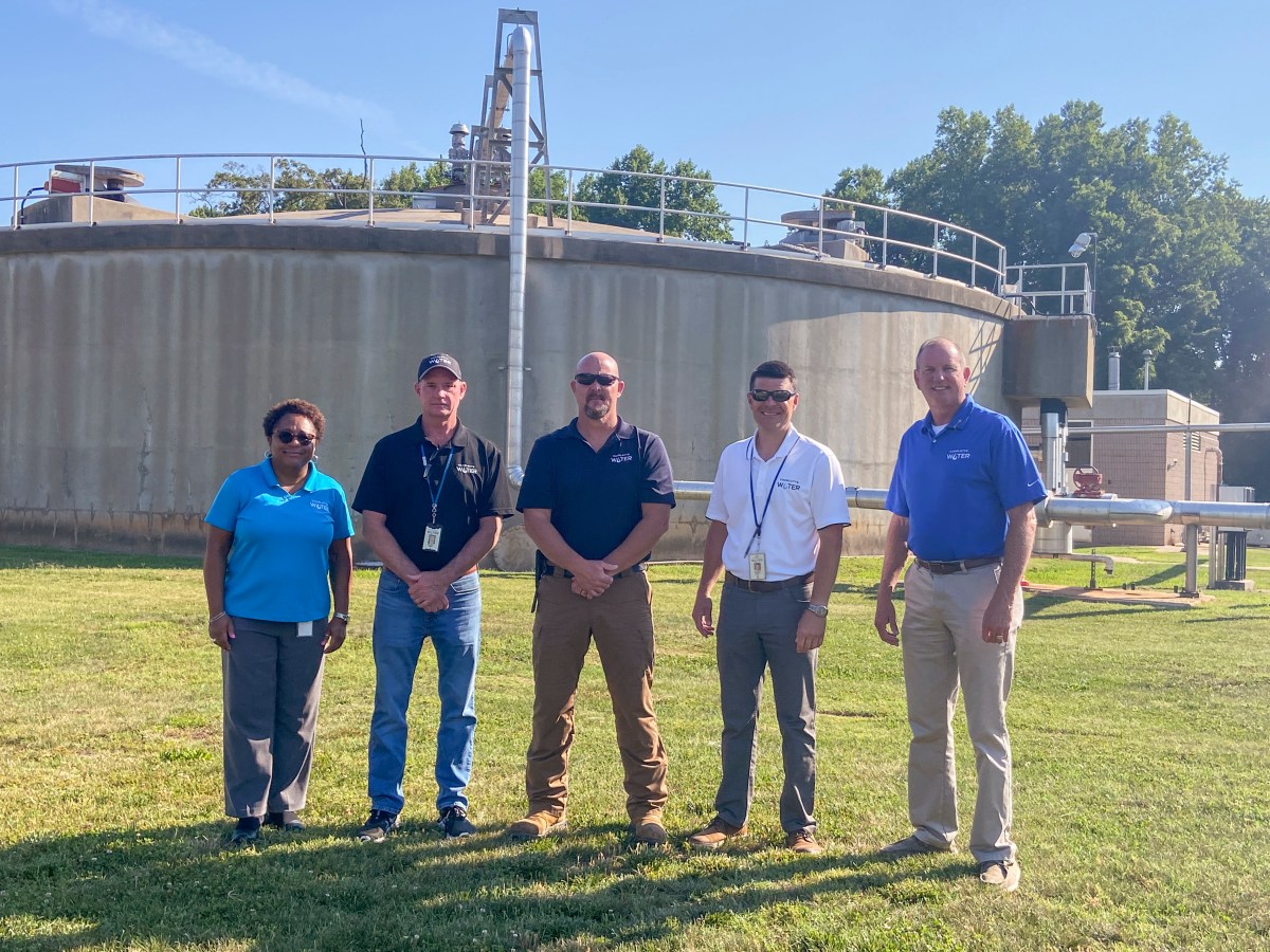 5 utility supervisors dressed in polo shirts stand on green grass in front of a large, round concrete water treatment structure