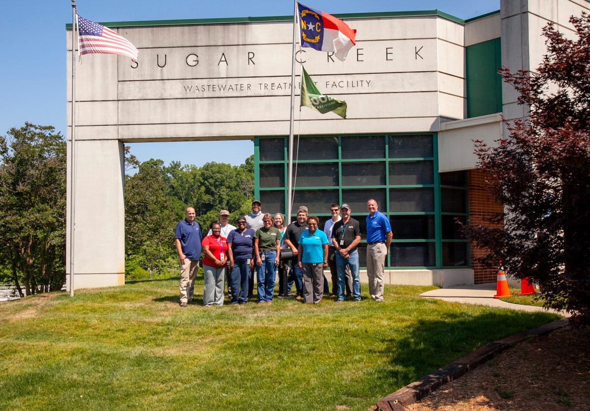 12 utility technicians and supervisors in polo shirts pose on green grass for a group photo in front of a large two-story concrete and green windowed building with the name Sugar Creek Wastewater Treatment Facility above
