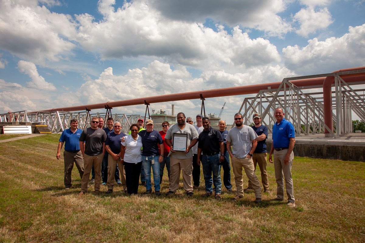 16 utility employees and supervisors line up on grass for a group photo, man in middle wearing a grey polo shirt holds a framed award plaque and behind the group is a large orange pipe crossing above them spanning across grey metal pipe supports