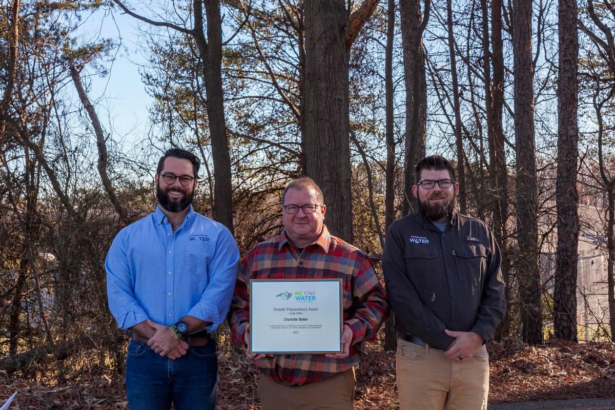 3 utility technicians dress in office casual wear pose in front of a wooded area, the middle person in a plaid shirt holds a wood and metal awards plaque