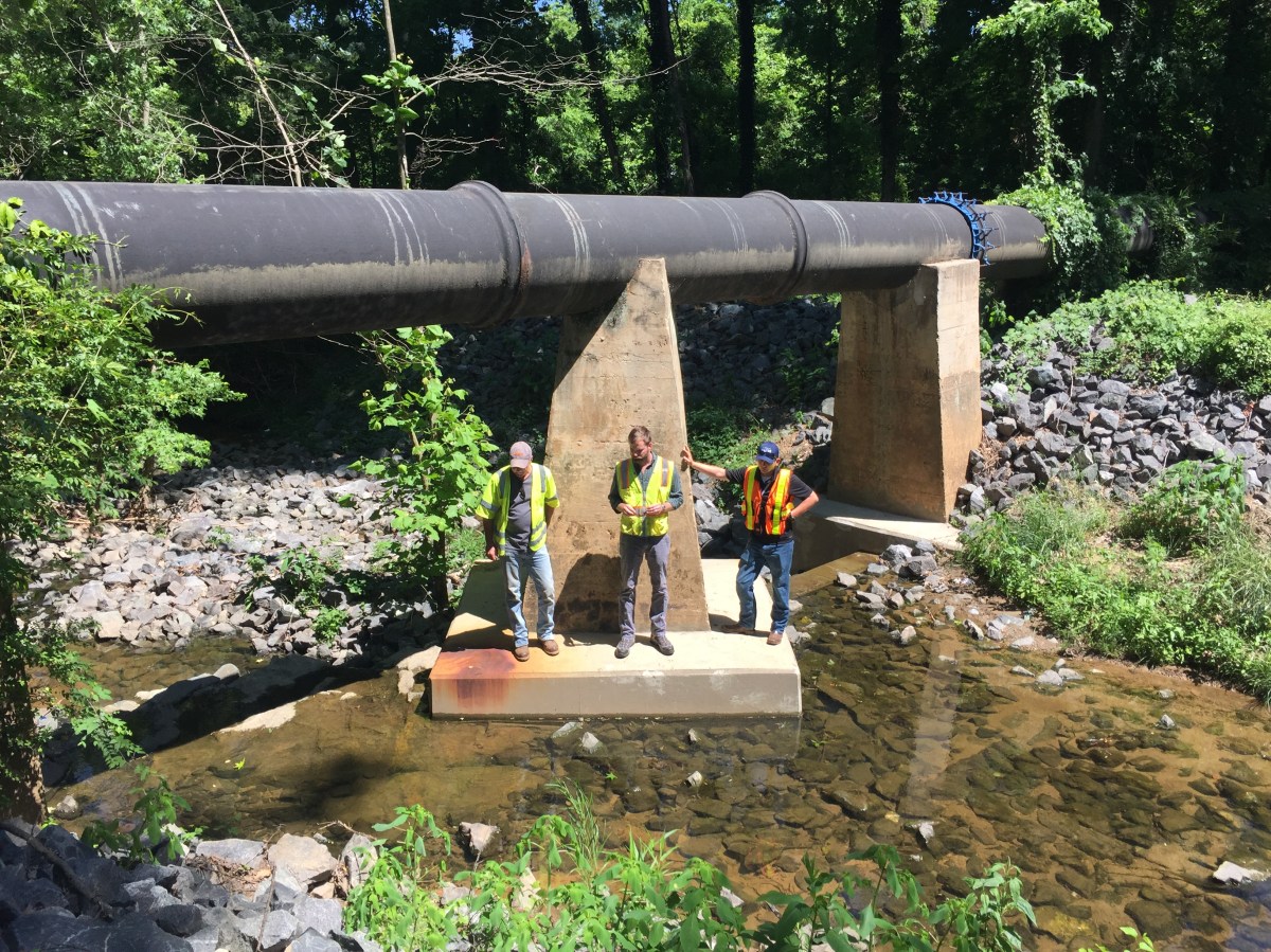3 utility technicians wearing yellow and orange safety vests, stand on the concrete dolphin of a large concrete piling that help supports a very large, brown steel pipe as it crosses over a rock littered creek
