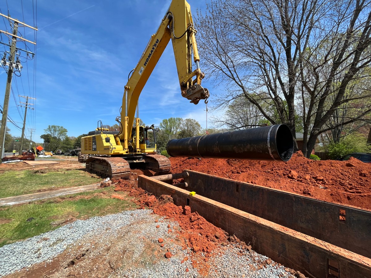 large excavator with a hook and cable lowers a large section of black pipe into a large, long brown metal box that has been installed in a large trench cut through the red earth