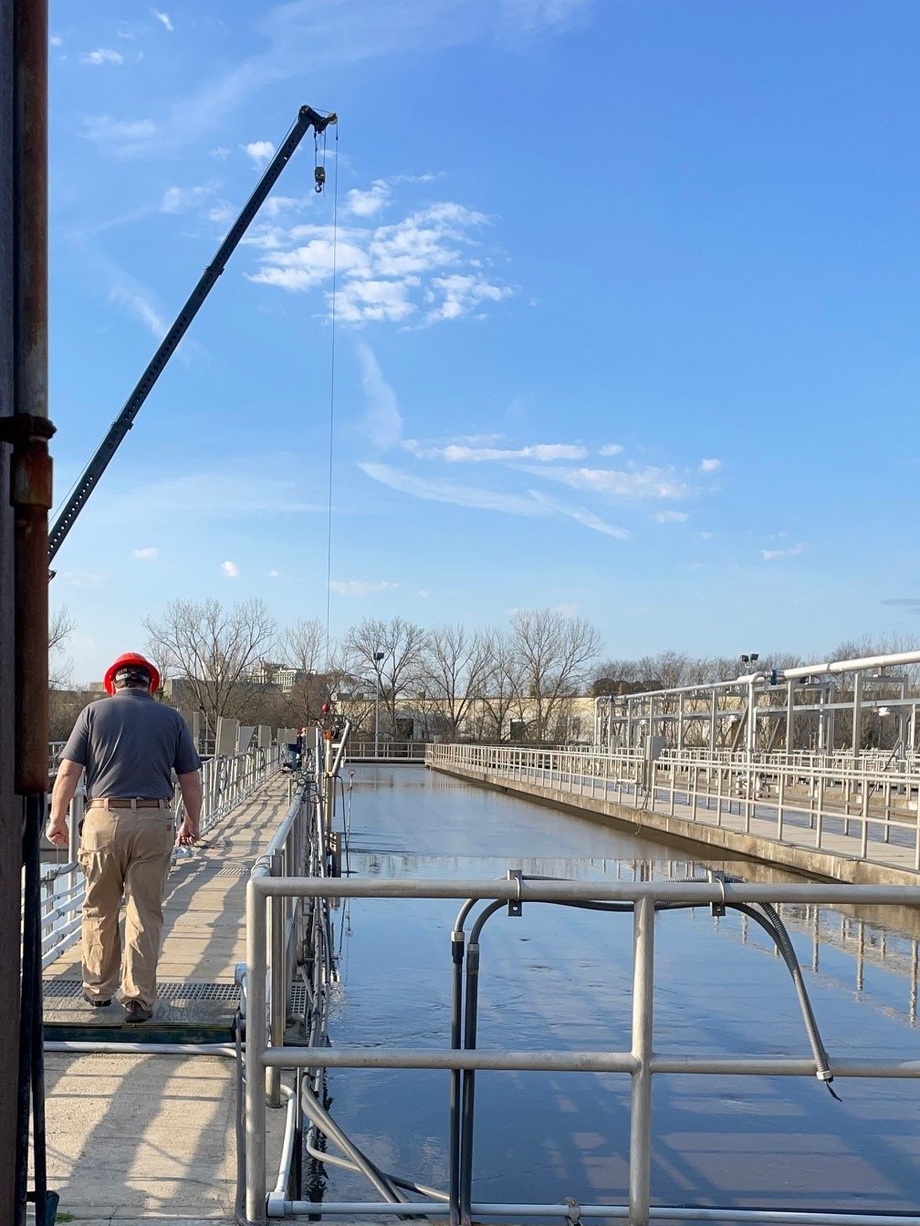 utility technician wearing an orange hard hat stands along a metal fence lined concrete causeway adjacent to a long narrow channel of water