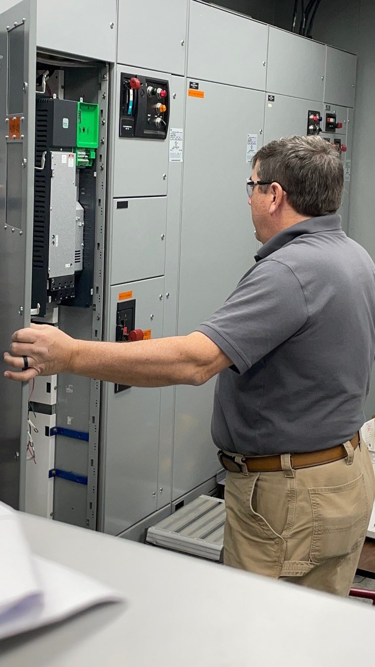 man in grey polo shirt and tan pants hold a tall, grey metal utility panel door open as he reviews the grey, green and black equipment inside