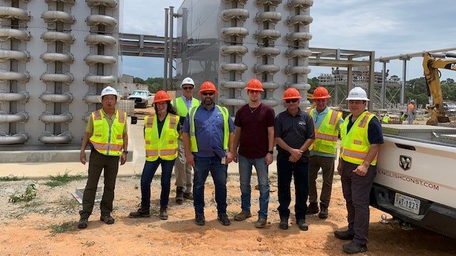 8 utility engineers wearing yellow safety vests and hard hats line up to pose for a group photo, they stand on dirt in front of a very large grey water treatment facility with lots of curved sections of pipes visible
