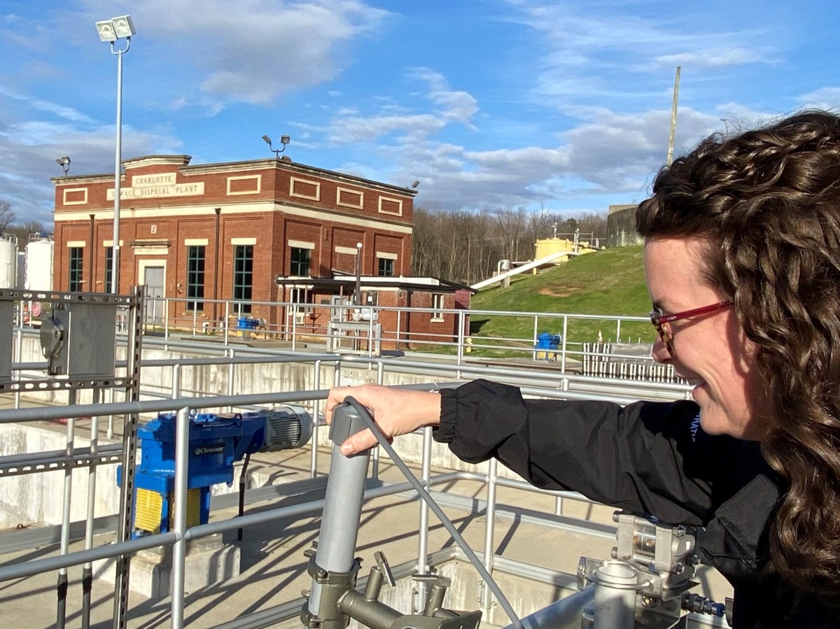 utility technician adjusts a grey cap on a short grey pipe with her hand as she overlooks a series of water treatment facility grey pipes and blue valves next to a one-story brick waste water disposal plant building