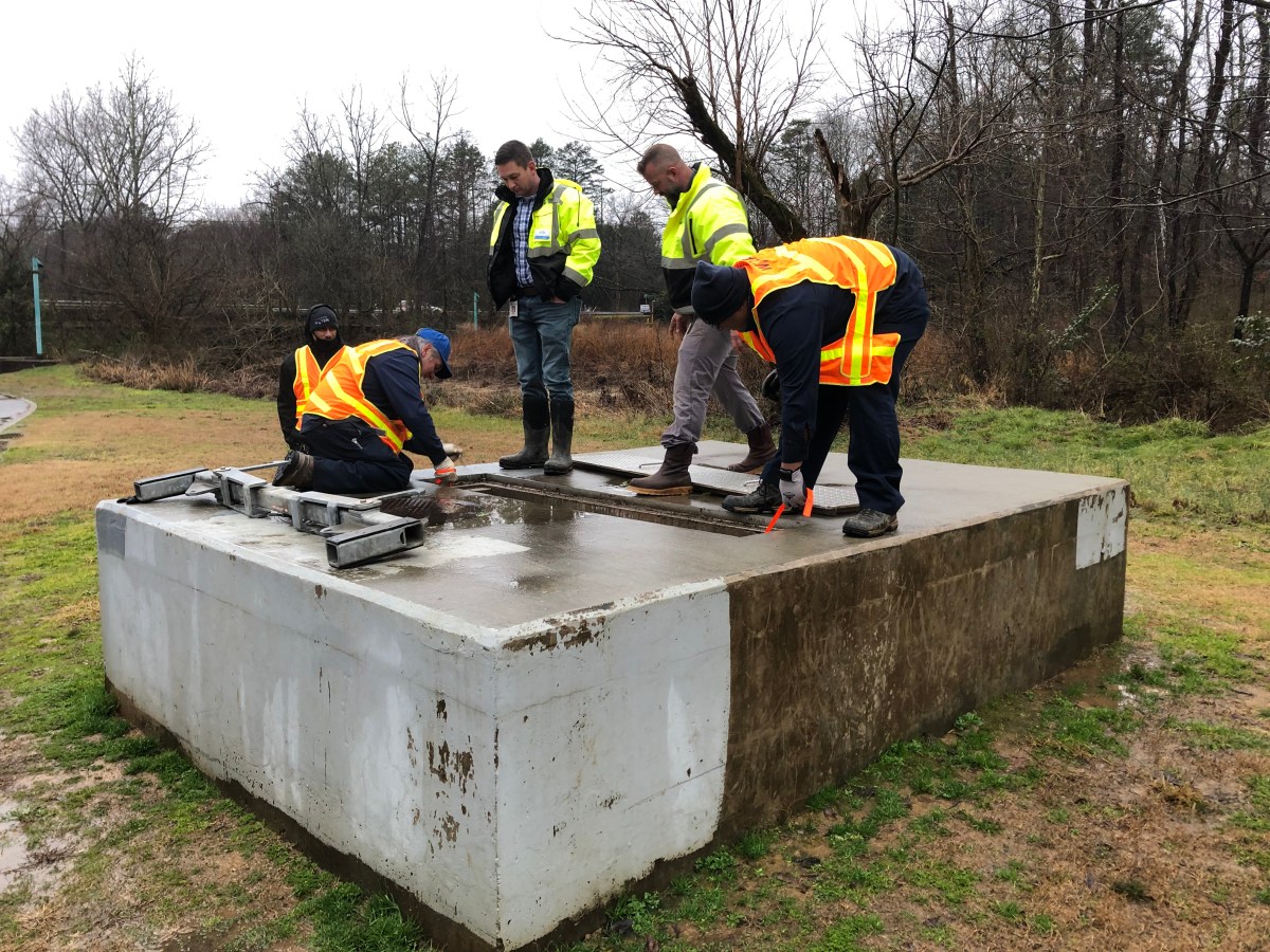 5 utility technicians wearing orange and yellow safety jackets clamber atop of a large rectangular concrete platform, they have removed a long thin rectangular panel to inspect what is inside and below