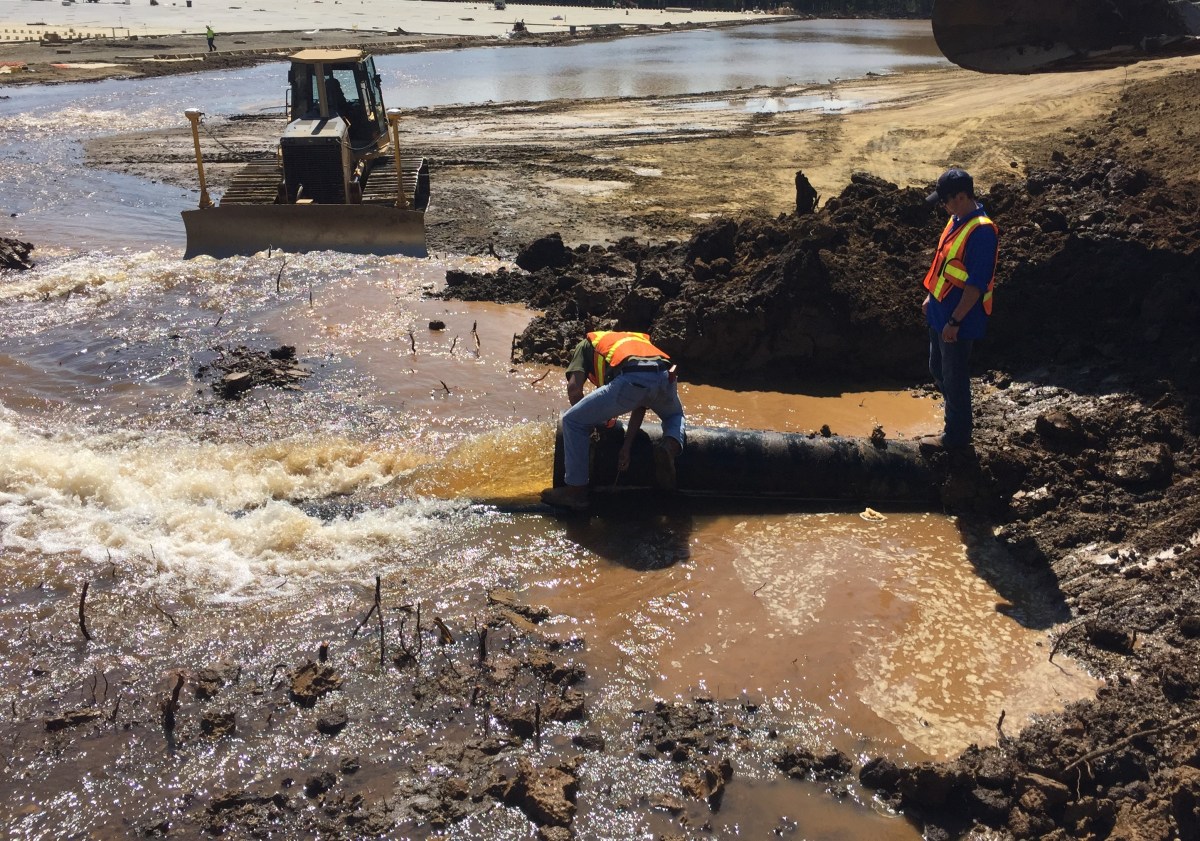 at a muddy job site, a utility technician wearing an orange safety vest crouches over a large brown pipe as run off water pours out into a shallow waterway, another utility technician wearing an orange safety vest stand on the pipe behind him and nearby a yellow bulldozer sits helping divert water flow