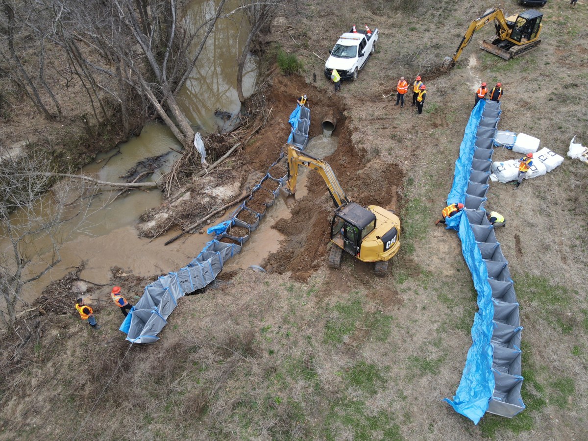 8 utility technicians stand near a yellow CAT excavator digs dirt at a muddy job site to divert running water run off from a protruding, underground black pipe, disturbed soil is put into a row of square blue holding bags that act as sandbags for the water diversion, a second row of blue square bags sit nearby