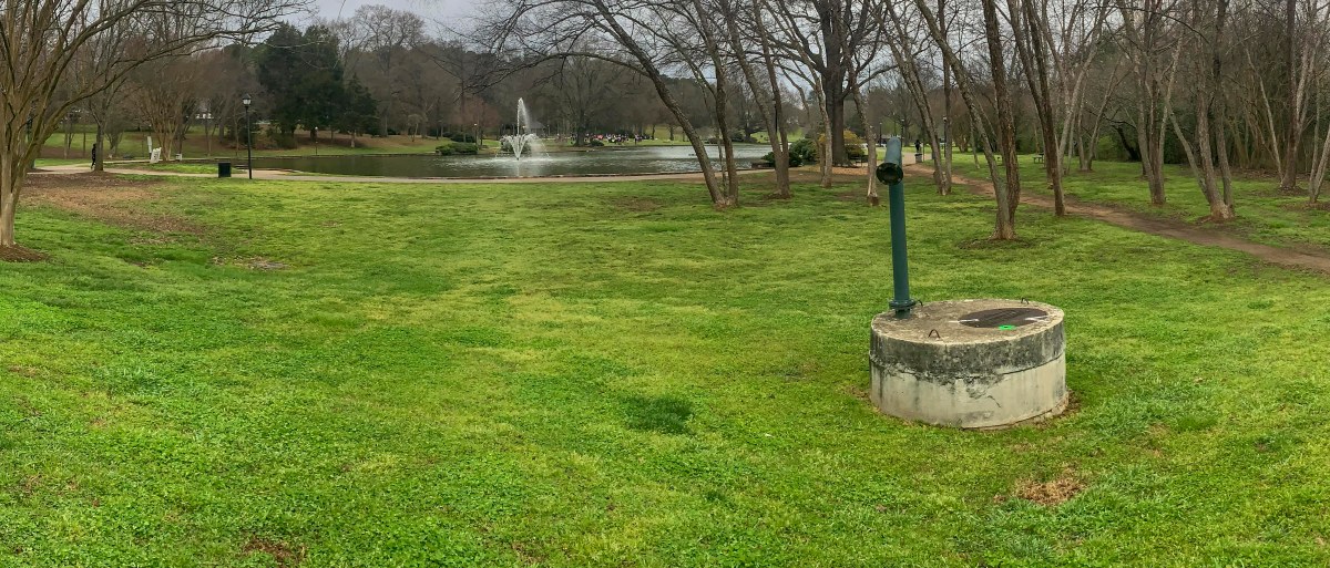 Round concrete sewer access platform with dark green pipe sticking out of it sits in the middle of a grassy, green park area with a pond an fountain in the distance