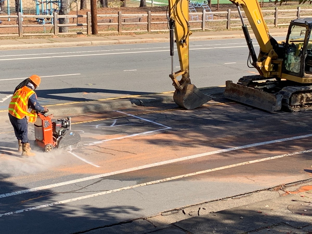 large yellow excavator is parked on the side of a street next to a utility technician wearing an orange safety vest and orange hard hat guiding an orange macadam circular to cut along the white lines painted on the side of the roadway