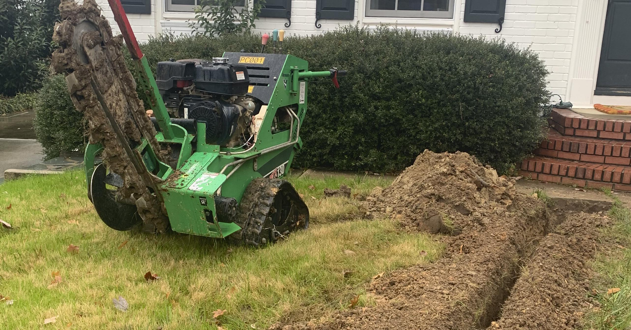 green, track driven ditch-witch sits on grassy lawn next to dirt trench dug in front of brick stairs at a white brick residence