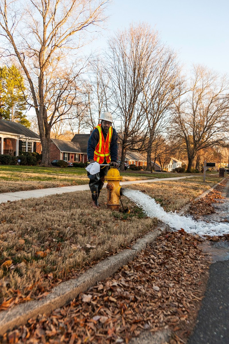 utility technician wearing orange safety vest and white hard hat, standing on a residential street, uses a large wrench to open the top valve on a yellow fire hydrant spewing water into the gutter filled with brown, autumn leaves