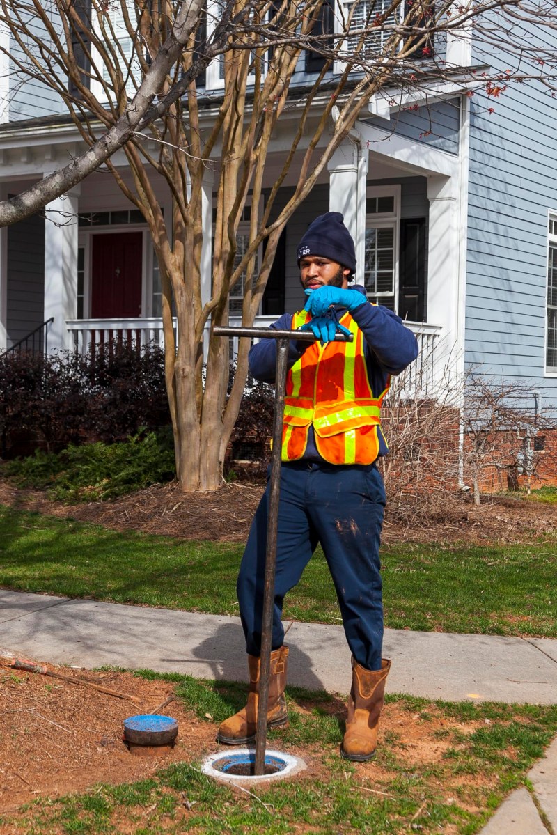 utility technician wearing a woolen blue hat and orange safety vest stands in front of a blue two-story house as he turns a large, T-shaped key inserted into a round hole in front of him, the blue metal lid for the hole sits nearby
