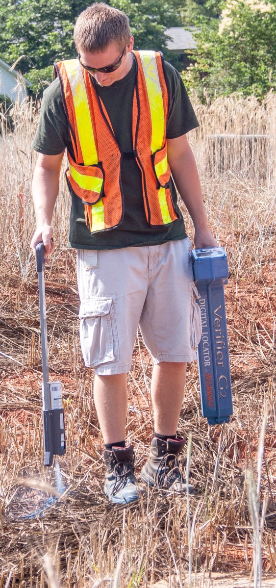 utility technician in tan shorts and black t-shirt and orange safety vest walks through a meadow of brown grasses holding a blue GPS tracking device and a long white and black paint sprayer to identify and mark a pipe leak underground