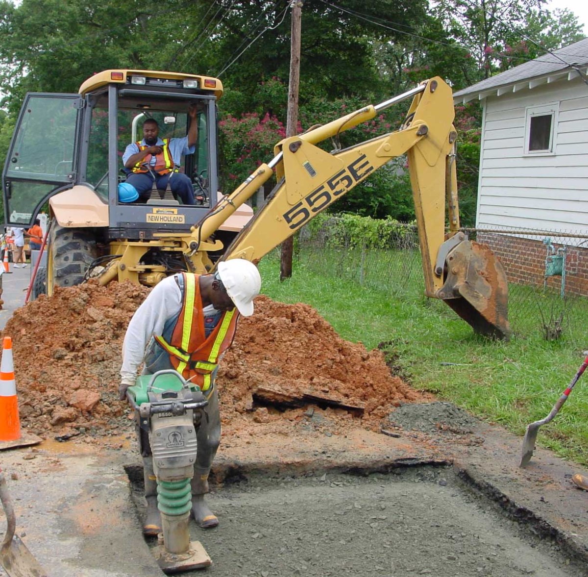 yellow 555E backhoe pauses streetside from refilling a large square hole with red earth and waits for a utility technician wearing an orange safety vest and white hard hat to finish tamping down the grey gravel soil in the hole with a light green, handheld power tamper.