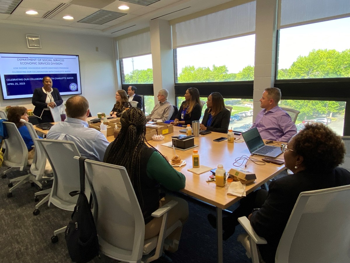 10 office worker seated in a white conference room with large windows, listen to a presentation by a woman in a navy blue suit and white blouse who stands at the head of the wood table, behind her is a large video screen stating Department of Social Services Economic Services Division Low Income Household Water Assistance Program celebrating our collaboration with Charlotte Water