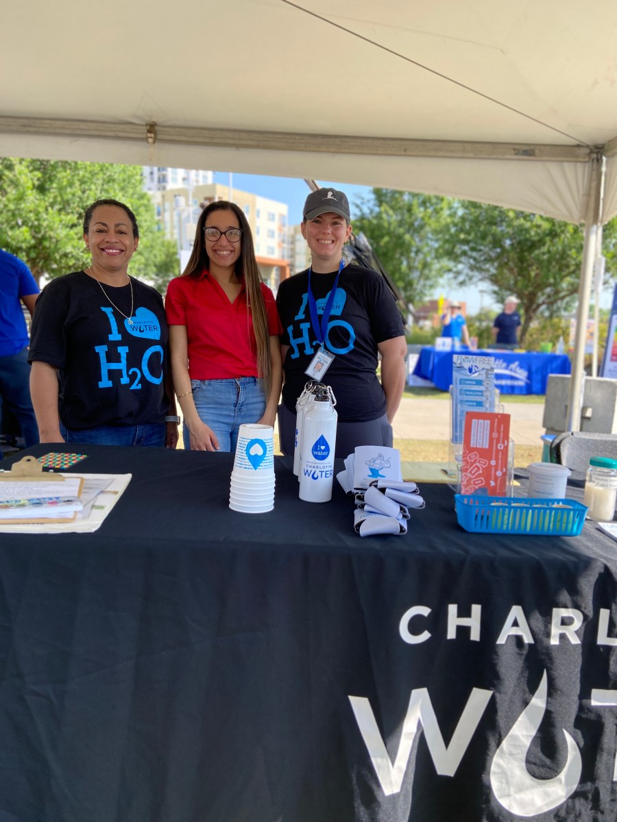 3 utility company employees, 2 navy blue t-shirts with light blue I heart h20 logos and one wearing a red polo shirt, stand behind a black table cloth with white Charlotte Water logo lettering, on top of table are several promotional items