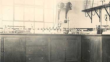 black and white photo of what an early water testing lab looked like with wooden cupboards topped with beakers, testubes, jars and other equipment