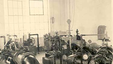 black and white photo of early water utility pipes, valves, and gages in a pumping station building