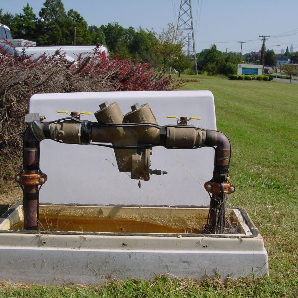 a large, white pvc case cover sits behind the utility company's backflow prevention pipe device, including brown metal valves and yellow levers, which emerges from the ground surrounding by a concrete and pvc base and green grass