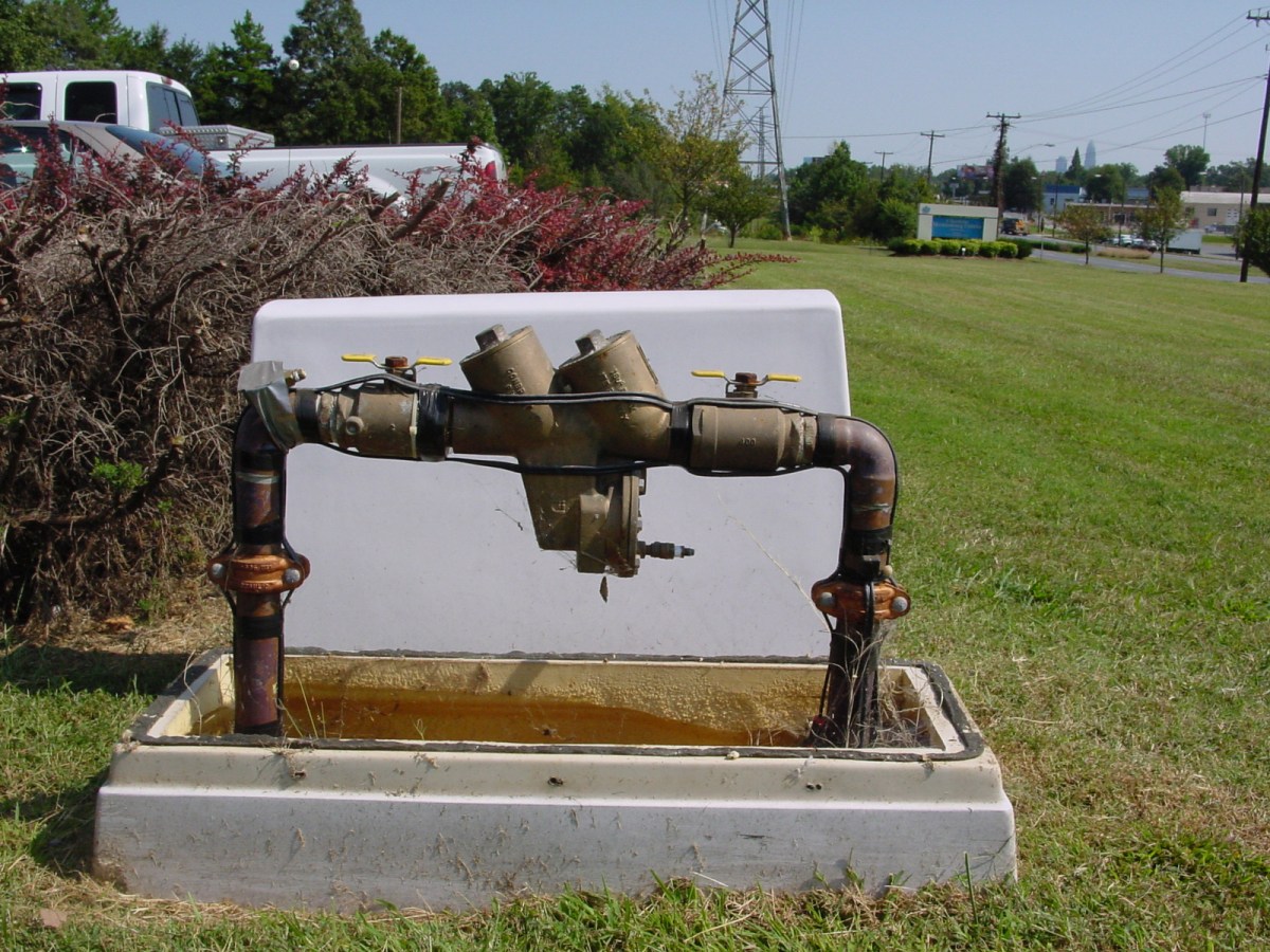 a large, white pvc case cover sits behind the utility company's backflow prevention pipe device, including brown metal valves and yellow levers, which emerges from the ground surrounding by a concrete and pvc base and green grass