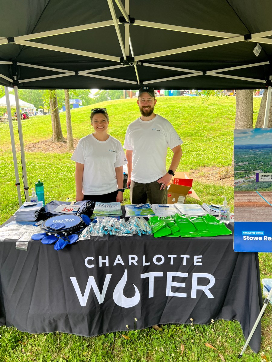 at an outdoor event, 2 employees wearing white t-shirts, one with black baseball hat, stand on grass underneath black tent behind table with black tablecloth with white lettering Charlotte Water; on top of table are many promotional items and brochures