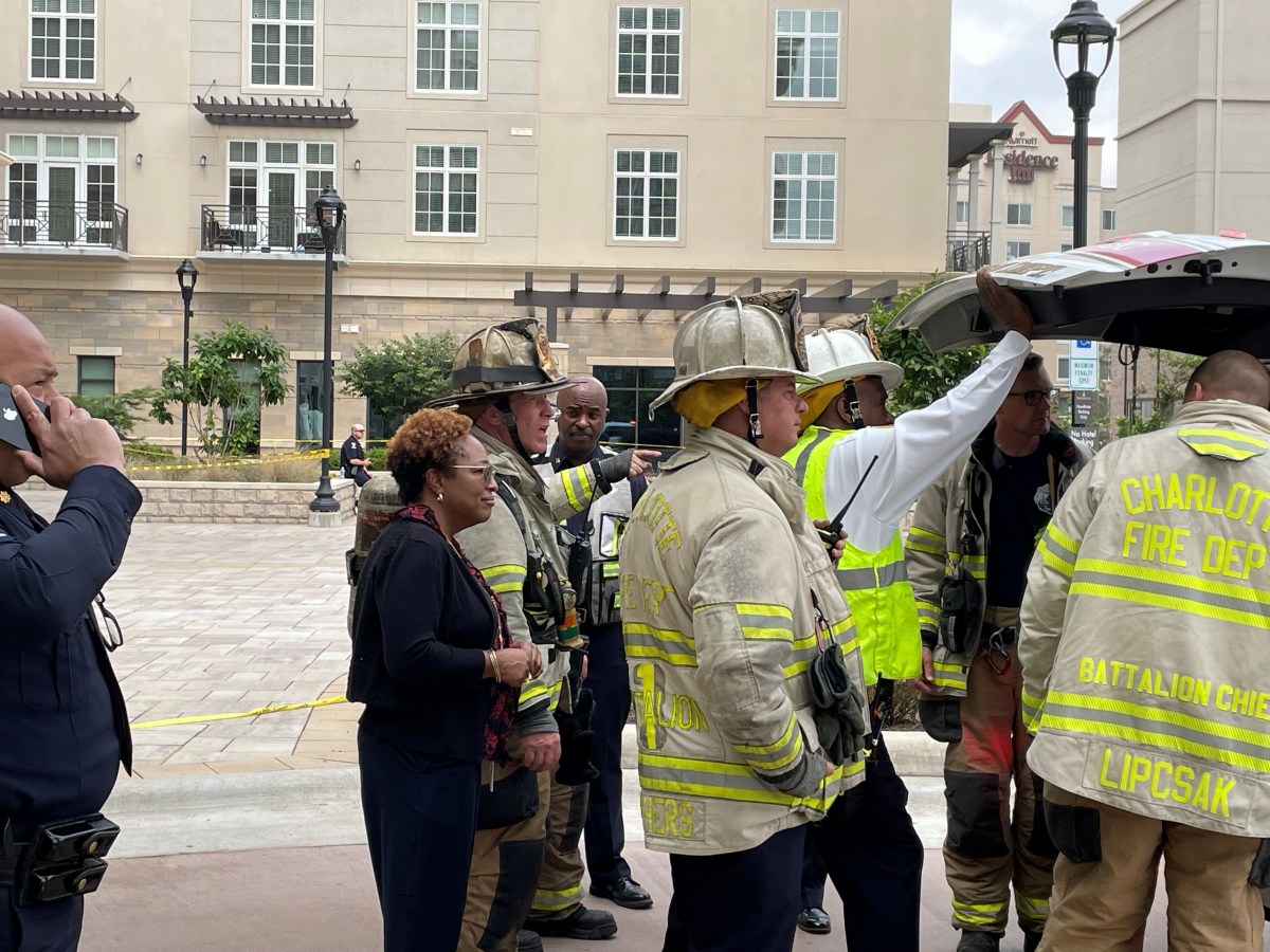 curbside on a city street, 3 utility company supervisors in office attire stand with 6 Charlotte Fire Department personnel dressed in their tan and yellow fireman's jackets and tan helmets, all looking into the open hatch of a white SUV