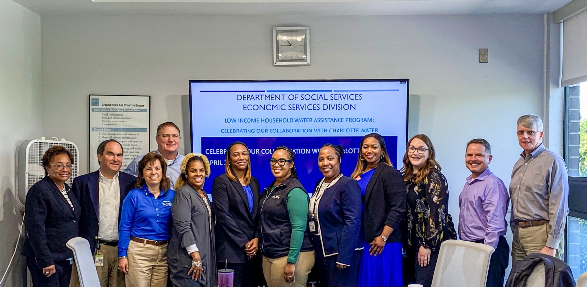 12 office workers line-up to pose for a group photo in a white conference room, behind them is a large video screen stating Department of Social Services Economic Services Division Low Income Household Water Assistance Program celebrating our collaboration with Charlotte Water