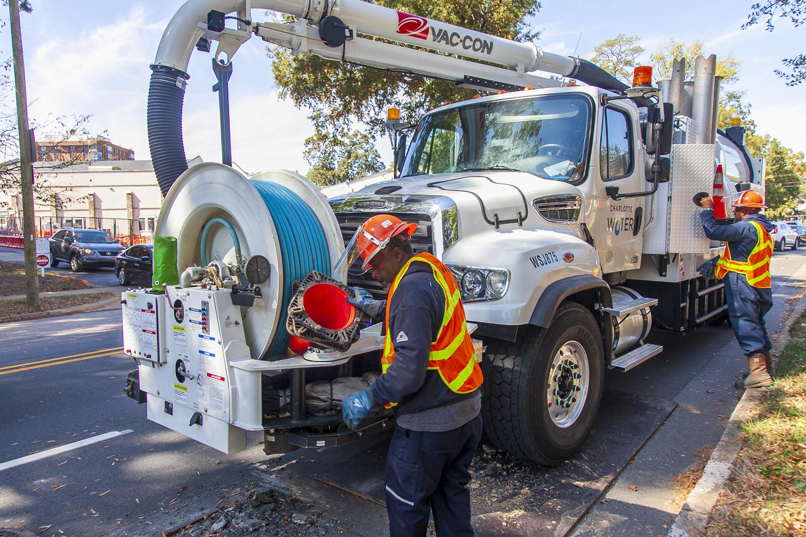 parked along a city street, a white Charlotte Water utility truck with septic capture tank and vacuum array including blue tubing on a giant spool, is prepared to be put to work by 2 utility technicians wearing orange safety vests and orange hardhats