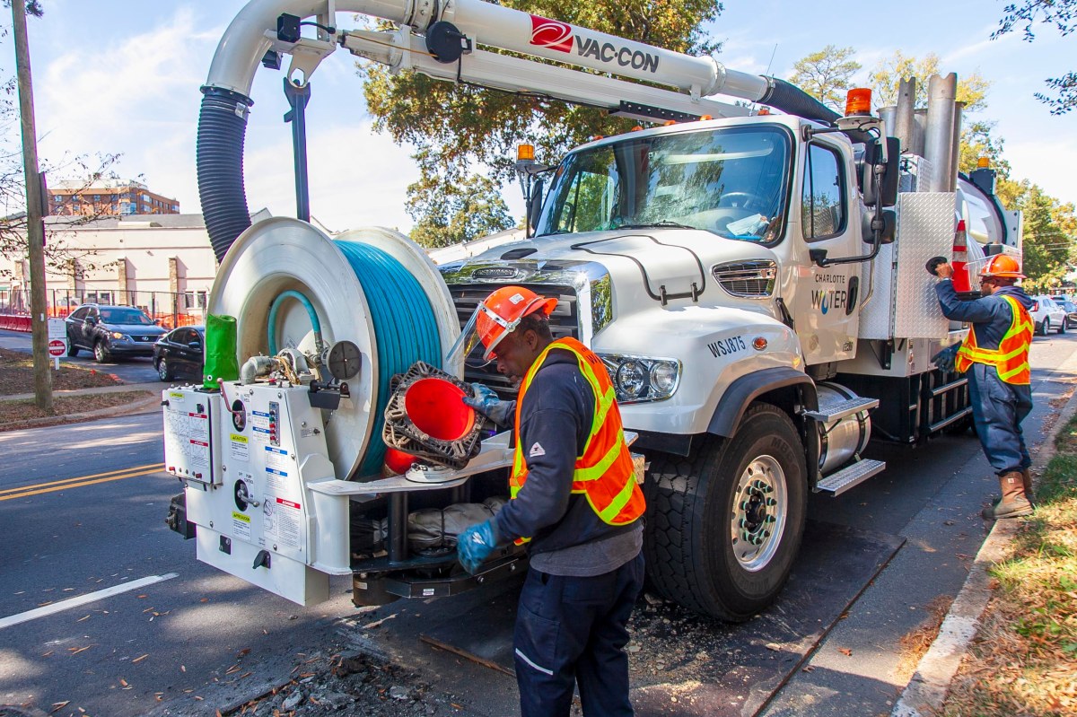 parked along a city street, a white Charlotte Water utility truck with septic capture tank and vacuum array including blue tubing on a giant spool, is prepared to be put to work by 2 utility technicians wearing orange safety vests and orange hardhats