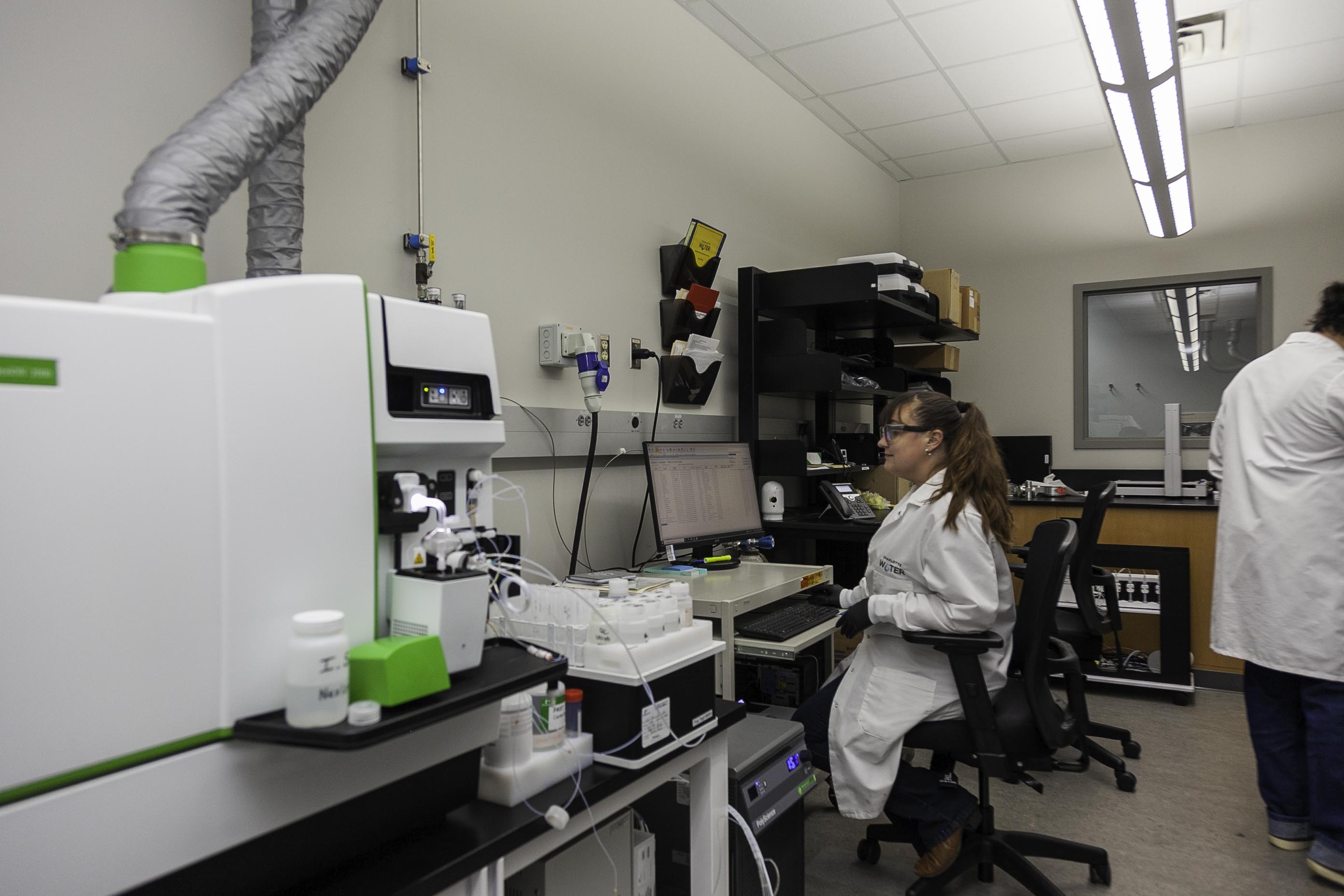 lab technician with long brown hair and white lab coat sits in black chair in lab, with lab equipment and machines surround her, she reviews data on a computer screen