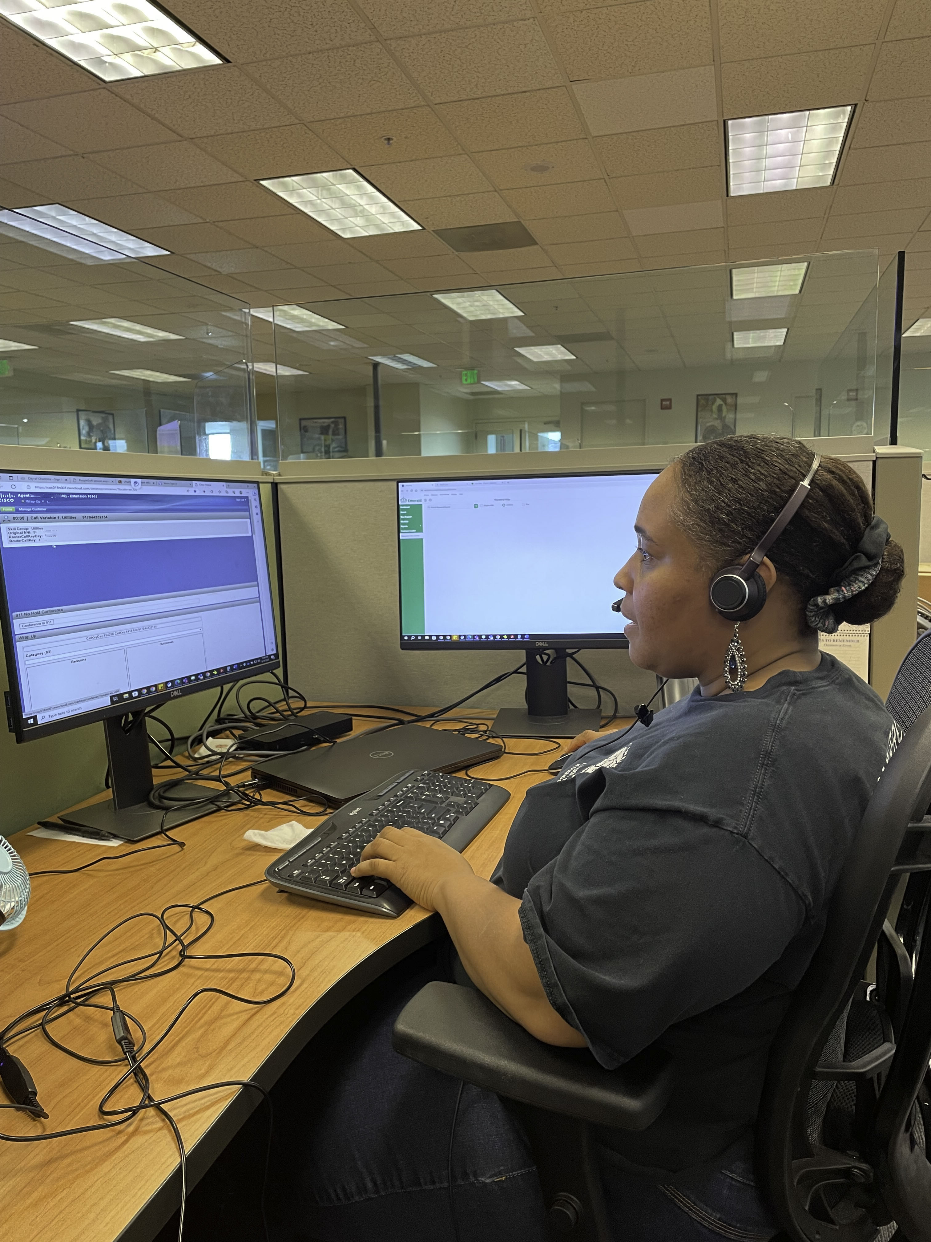 office worker seated at a wooden desktop wearing dark grey t-shirt and headset speaks to a caller while she reviews data on a large computer screen and types on a black keyboard