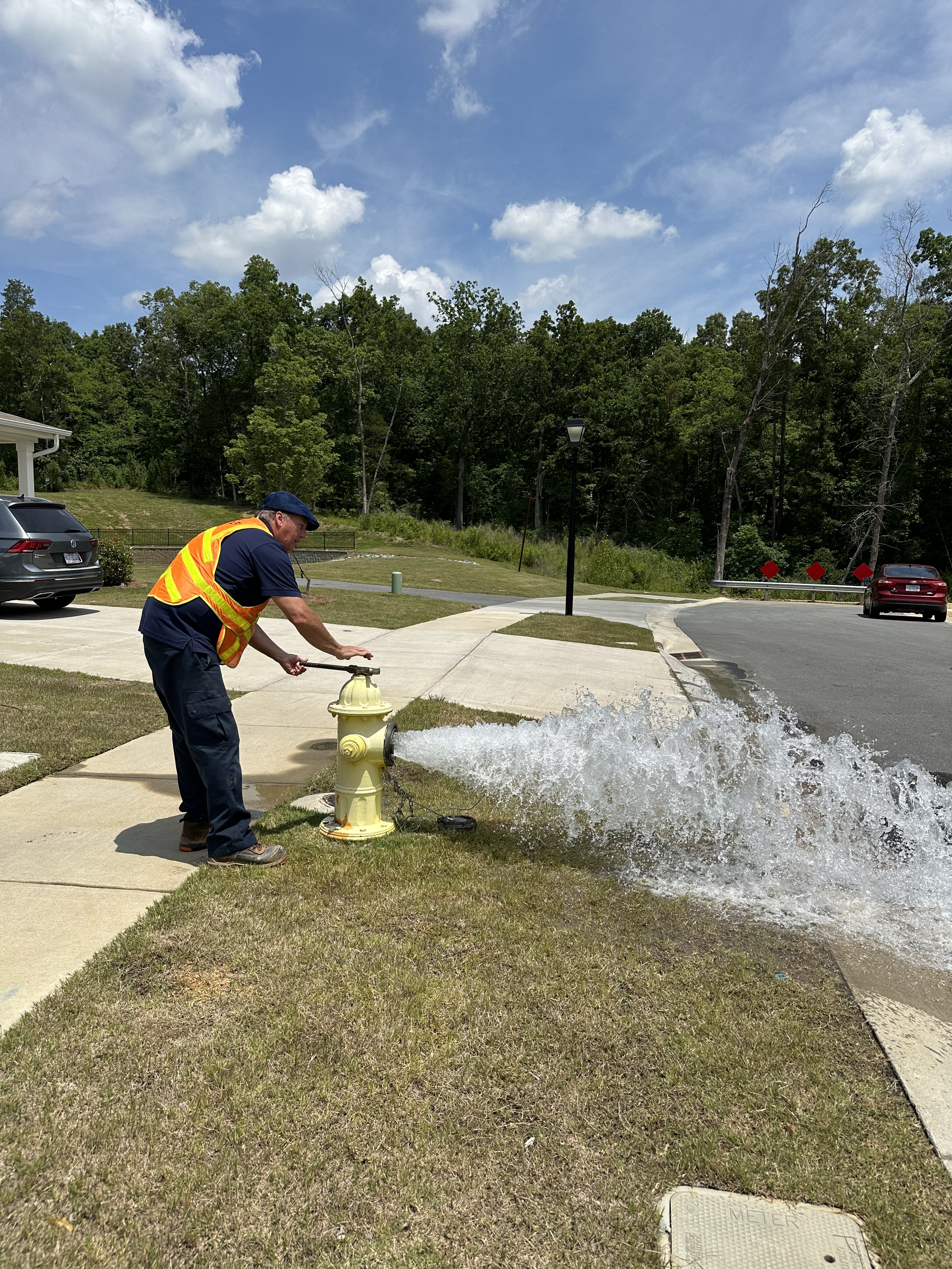 utility technicians wearing orang safety vest and dark green cap uses a large pipe wrench to open the valve on a yellow fire hydrant spewing water curbside on a suburban street