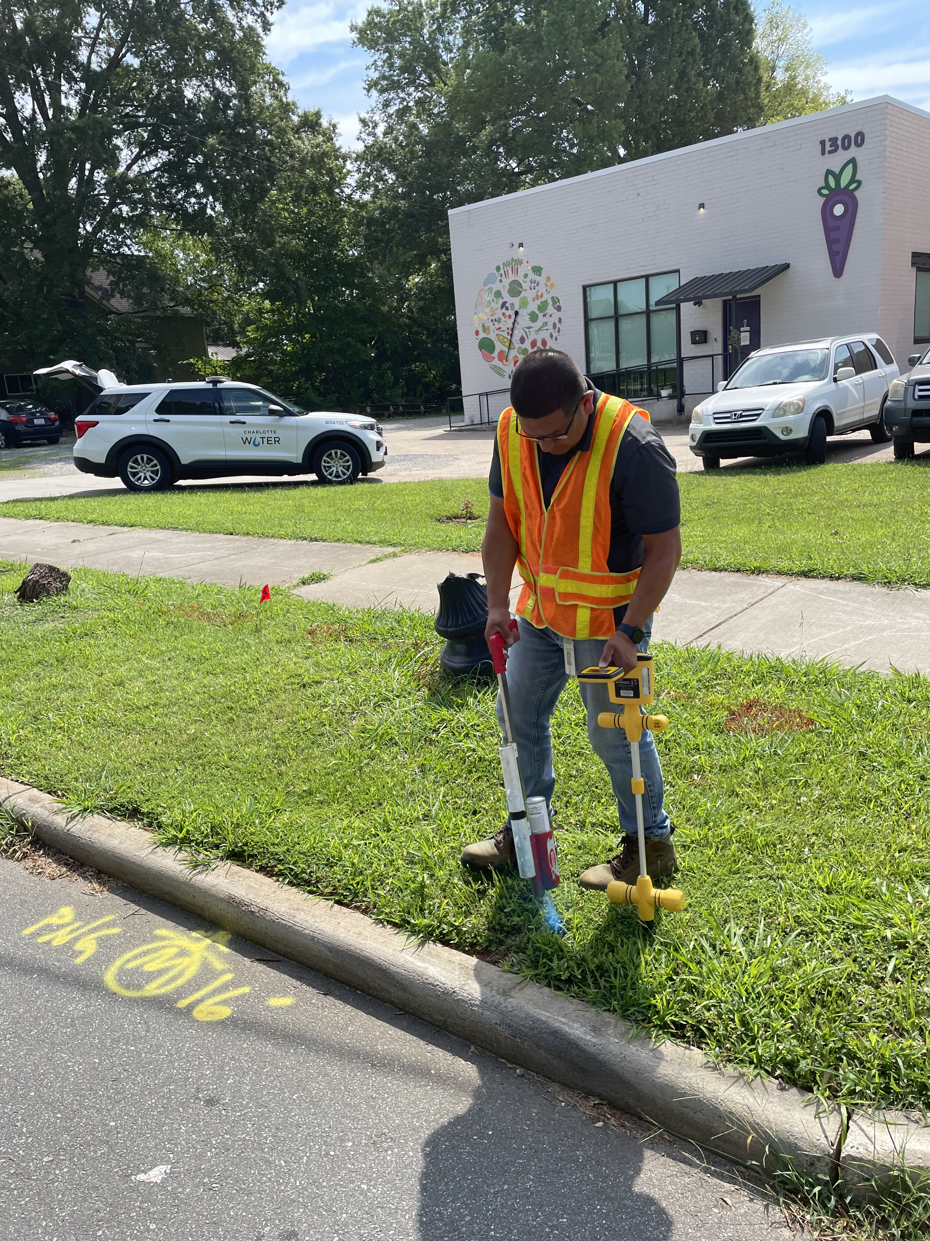 utility technician stands on grassy area between curb and sidewalk holding a yellow GPS mapping device and a red and white spray paint tool, he is checking where to mark a spot