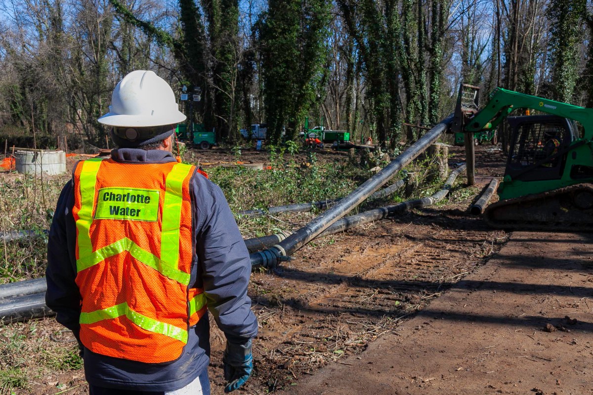 utility technician wearing orange safety vest with Charlotte Water labeling and white hard hat stands on dirt path in green forested area watching a small green skid loader lay a section of black pipe into place along the ground
