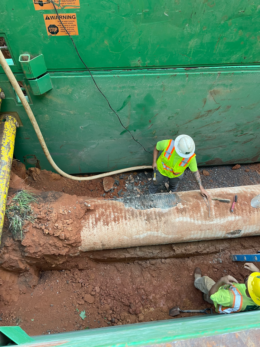2 utility technicians wearing yellow safety vests and hardhats inspect an excavated concrete sheathed pipe as it passed through red earth in a green metal chamber, the sheathing is crumbling away revealing grey metal