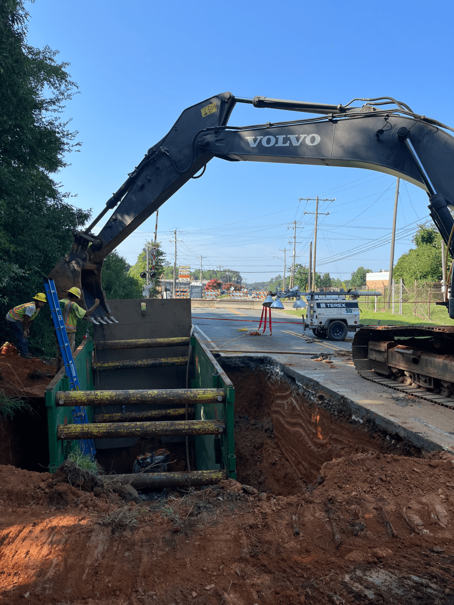 arm of a grey volvo backhoe's arm is poised above a green and yellow metal box that has been excavated along a roadway, 2 utility technicians, a pile of red earth and construction equipment are nearby