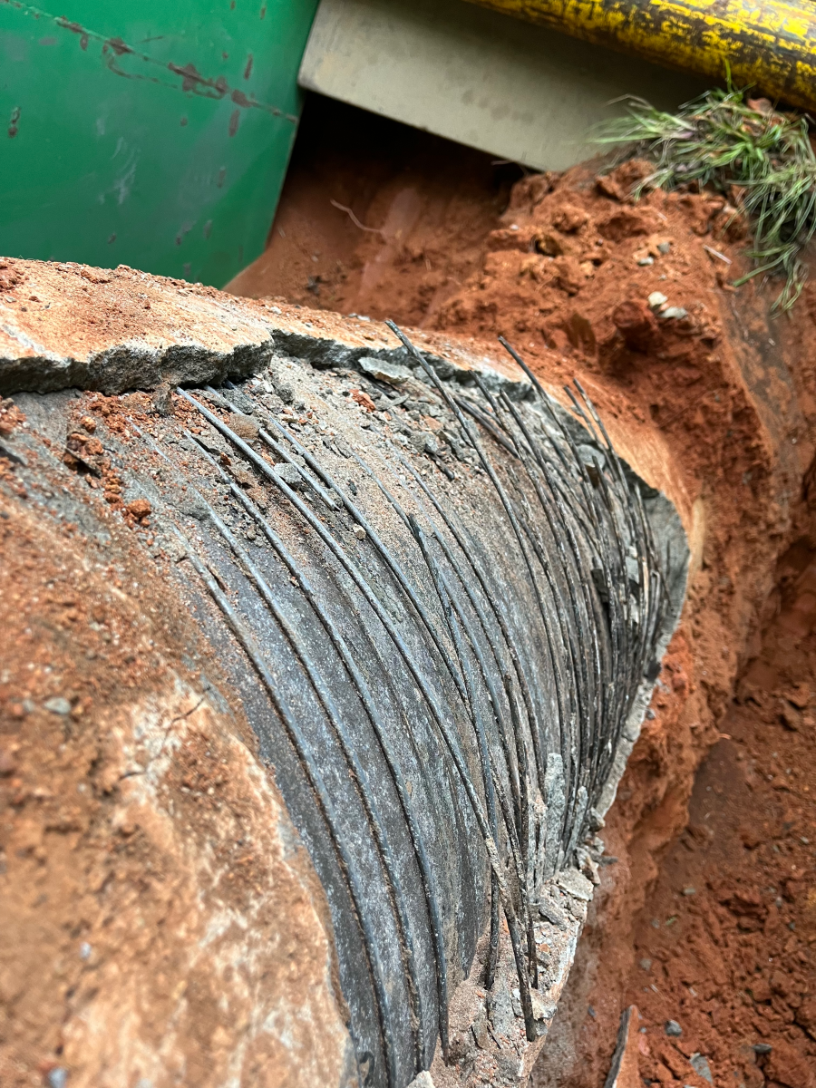 view of crumbling concrete sheath and broken rebar bands exposing a section of grey pipe as the red earth around it has been excavated