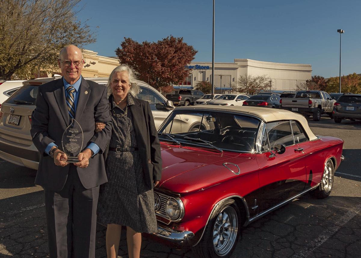 Man in grey suit holding a water droplet shaped glass award, he stands outdoors next to a woman in a black jacket and grey patterned dress and shiny, red 1960's convertible with a white top