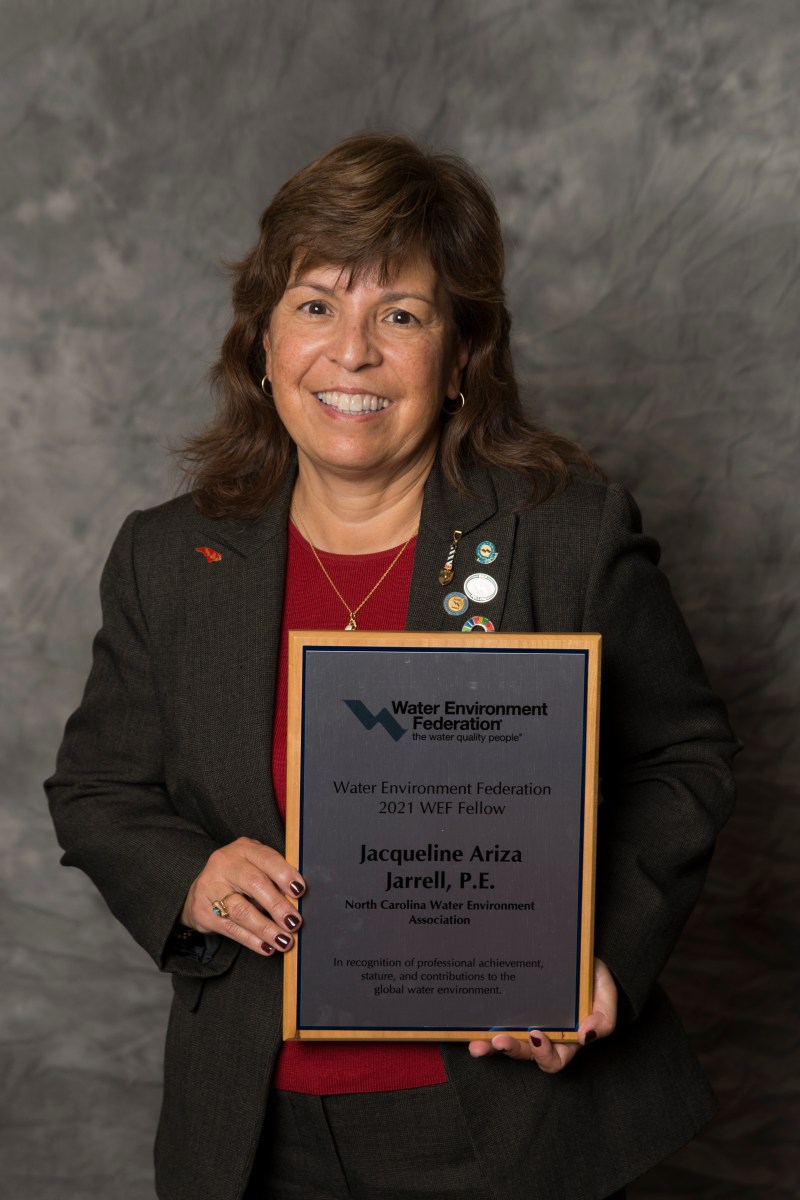 Woman with long brown hair wearing a red sweater and black business suit with round, colorful buttons on her lapel and holds a wood and inscribed metal plaque.