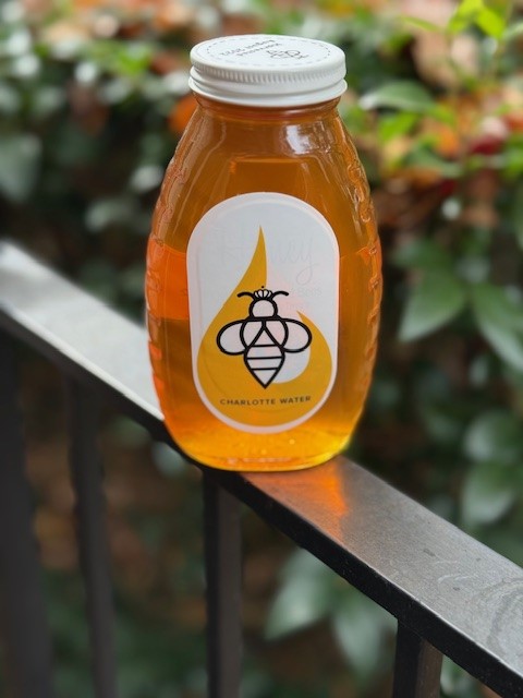 Sitting on a metal railing is a jar of golden honey with white lid and a yellow and white label with the Charlotte Water and honeybee logo on it