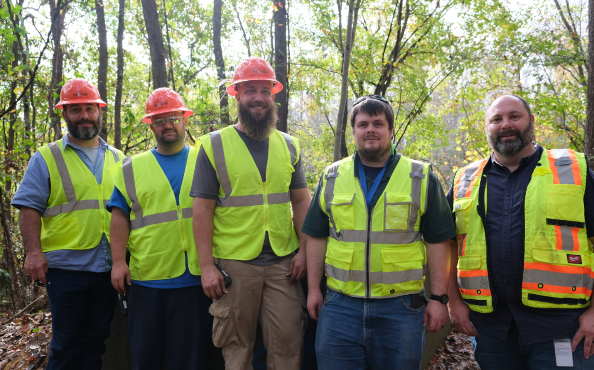 5 utility technicians wearing yellow safety vests and pose for a group photo, they stand in front of green, forested area