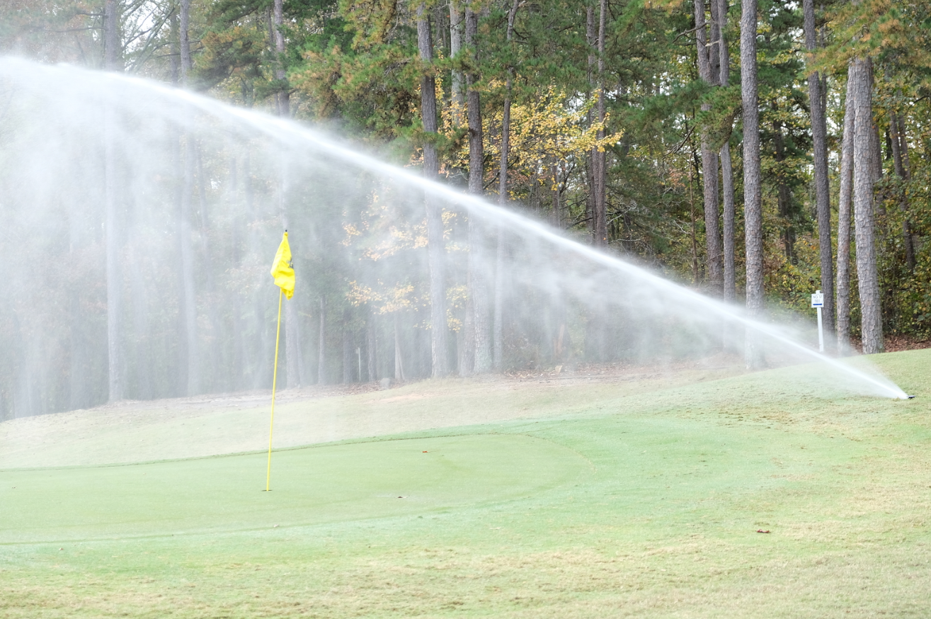 A lawn sprinkler sprays water across the green grass of a golf tee, a yellow tee flag appears behind the spray