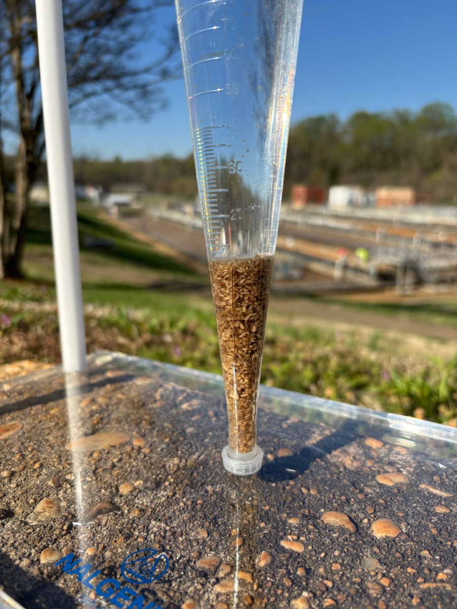 Clear glass measured rain gauge is filled with brown debris sites on a clear plexiglass lid over dirt