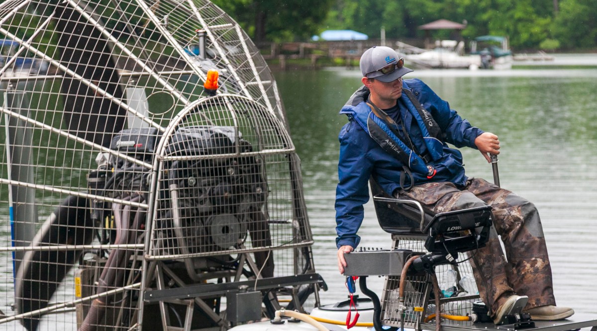 young man wearing blue windbreaker and grey ball cap sit in pilot's chair of a fan driven swamp boat on a lake