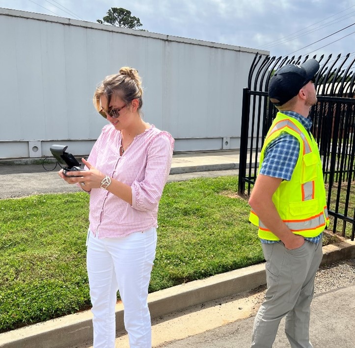 Student intern in pink blouse adjusts her camera while standing next to a utility employee in a yellow safety vest and a black iron fence