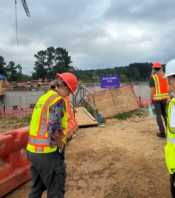 Student intern with patterned blue shirt and yellow safety vest pulls something out of her pocket as she tours a utility construction site with dirt, lumber, concrete and a sign building 910